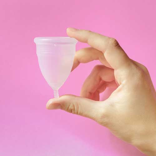 A hand holding a the menstrual cup on a pink background.