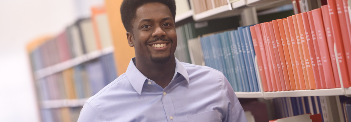 A male student standing in front of a row of library book shelfs.