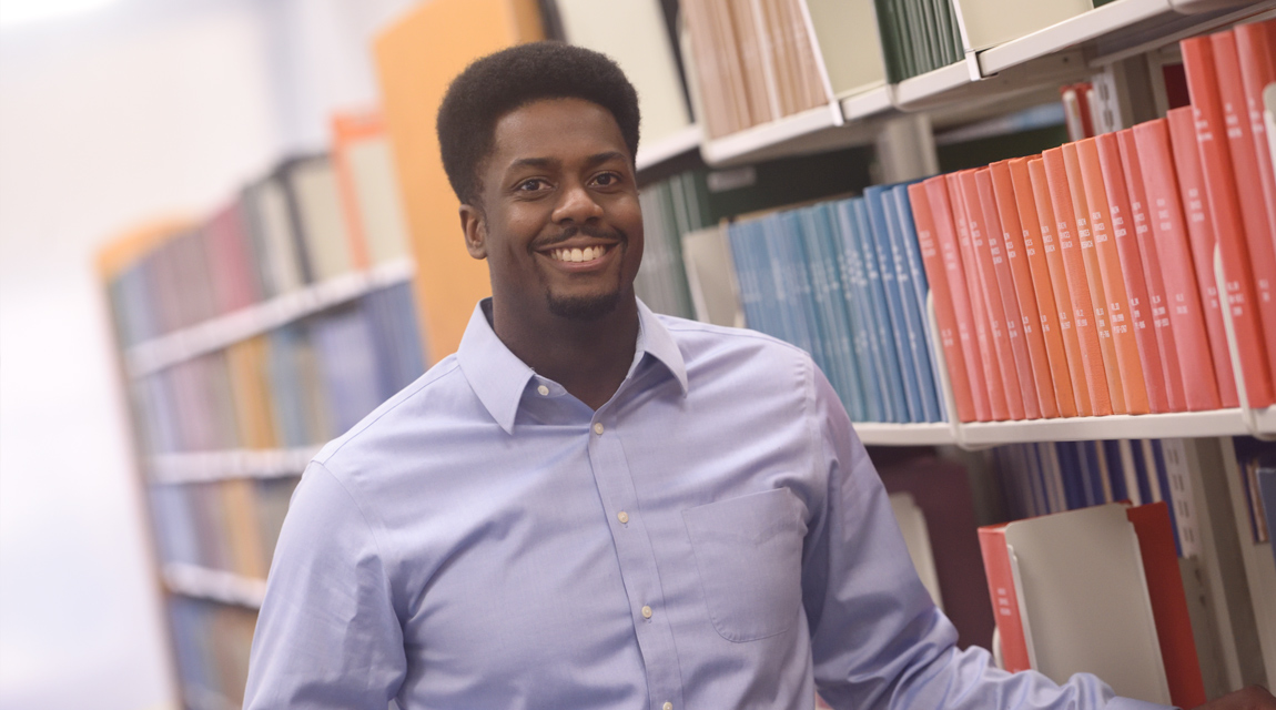 A male student standing in front of a row of library book shelfs.