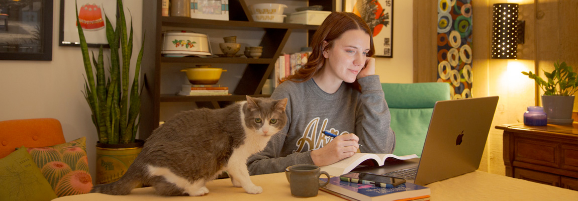 A student sitting at her dining room table looking at her computer. There are books on the table and her cat is providing company.