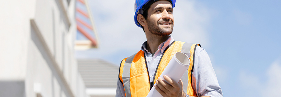 A man wearing a hard hat and a safety vest at a construction site. The man is also holding a roll of building plans.