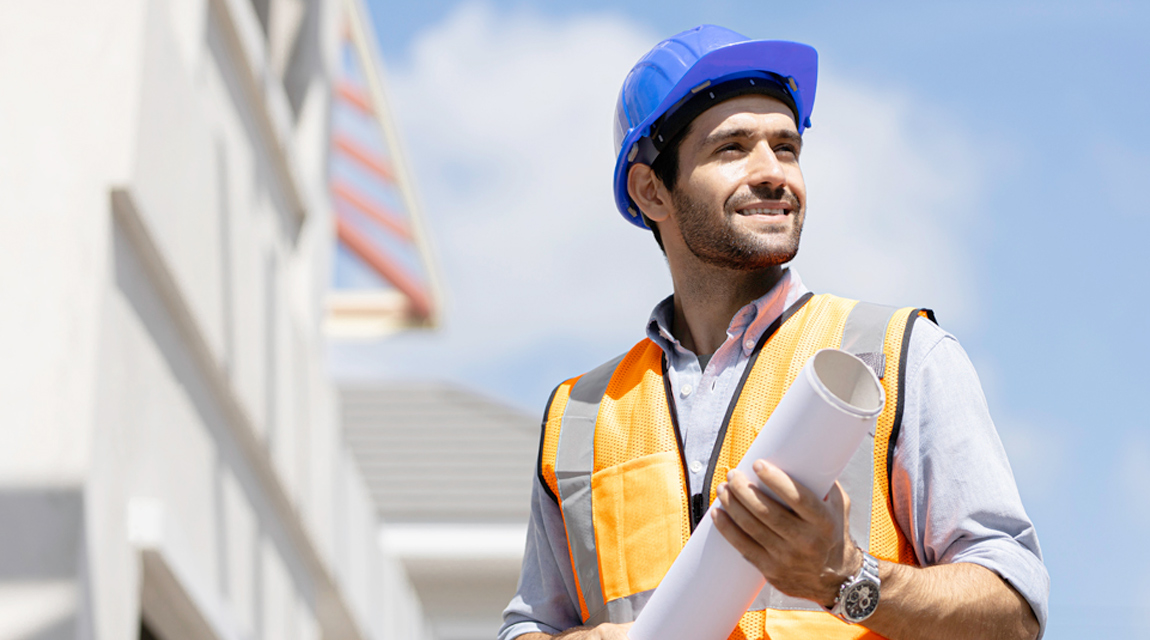 A man wearing a hard hat and a safety vest at a construction site. The man is also holding a roll of building plans.