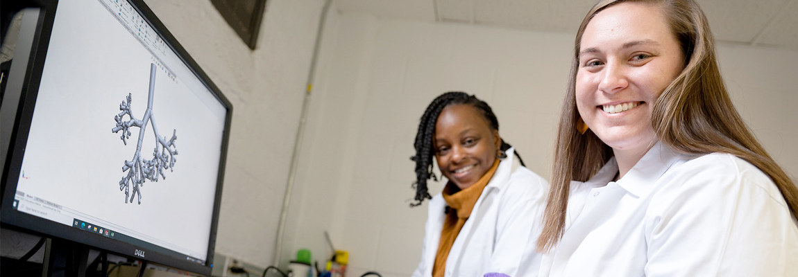 Two females wearing lab coats looking at the camera. Both are sitting in front of a monitor with scientific figure on the screen.