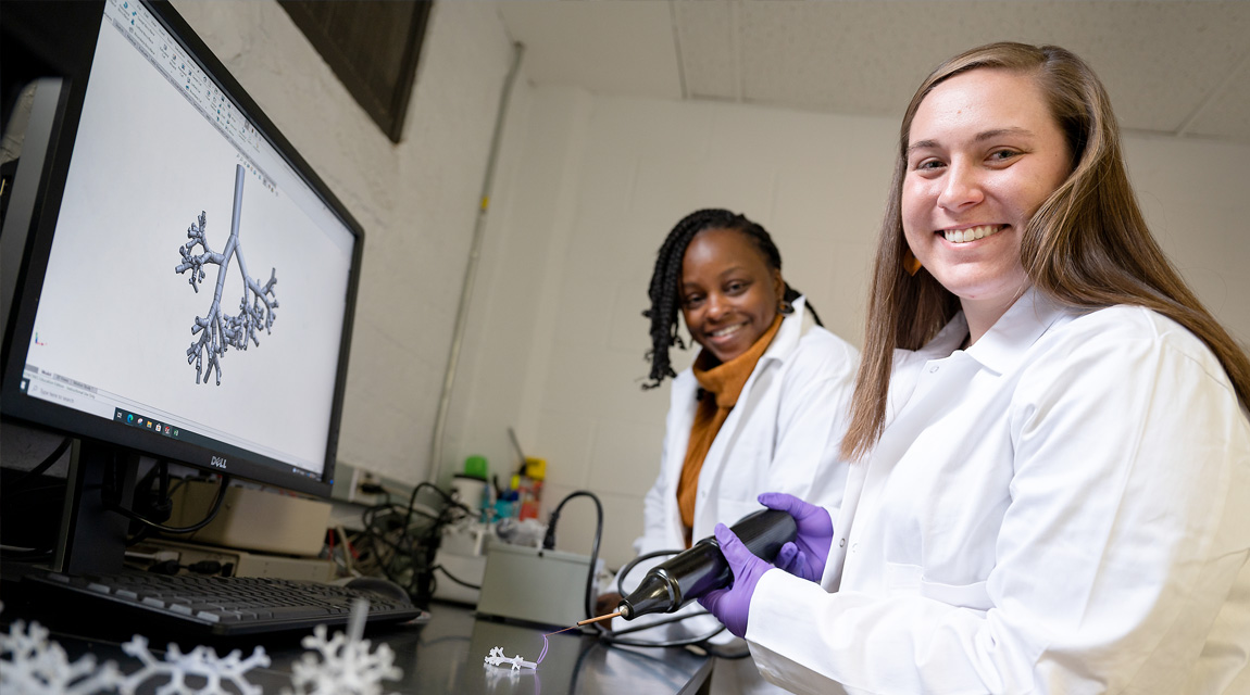 Two females wearing lab coats looking at the camera. Both are sitting in front of a monitor with scientific figure on the screen.