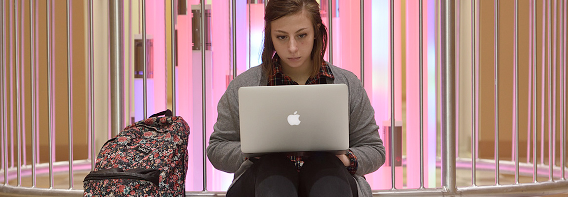 A student sitting on the floor with a laptop on her lap and a backpack next to her. She is facing the camera but looking at her computer.