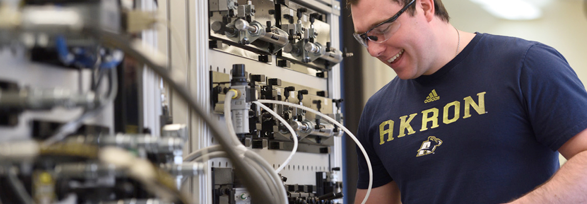 A smiling student standing in front of a wall of industrial controls.