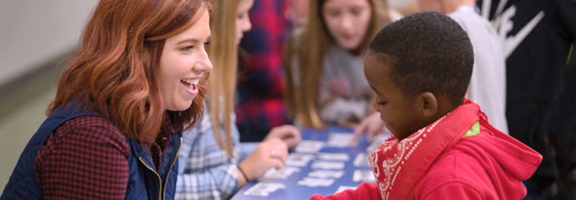 A female teacher talking with a young male student with other students in the background.