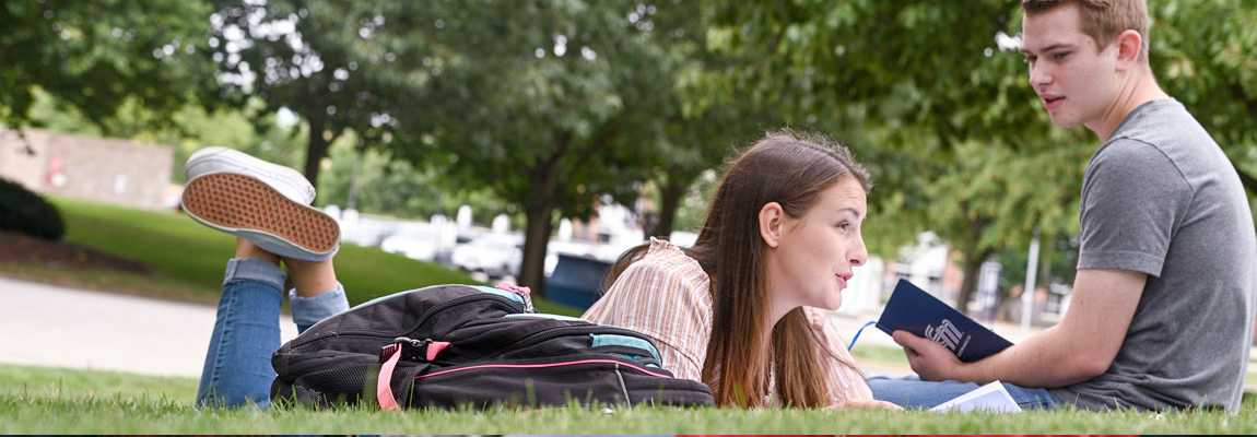 Two students talking in the grass. One is laying on her stomach with a book and the other is sitting on the grass.