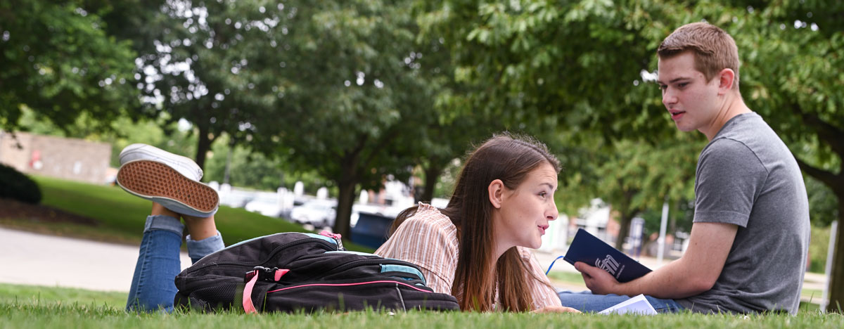 Two students talking in the grass. One is laying on her stomach with a book and the other is sitting on the grass.