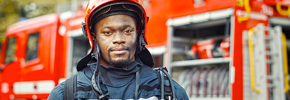 A male firefighter wearing a helmet and other safety gear standing in front of a fire truck.