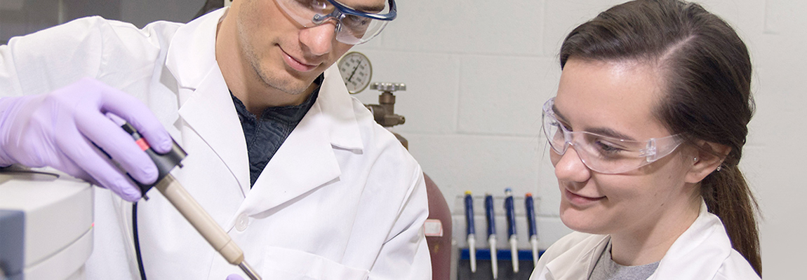 Two graduates students in a lab. Both students are wearing lab coats and goggles. The student on the right is looking to an instrument held by her co-student.