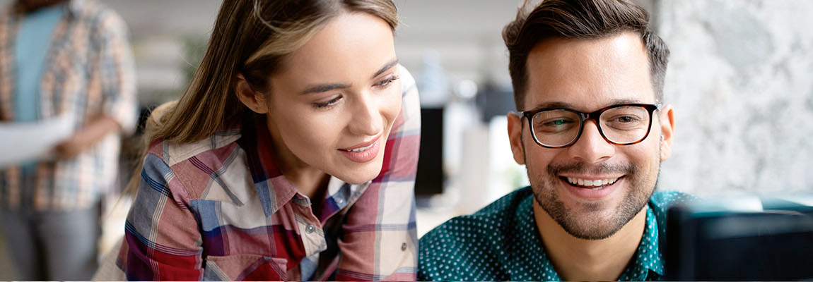 A young woman sitting at a computer with a young man standing behind her looking at the computer screen.