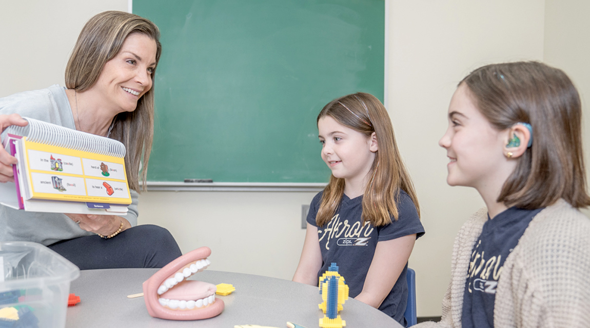 A speech pathologist sitting in a chair working with two young clients. The pathologist is holding a picture book.