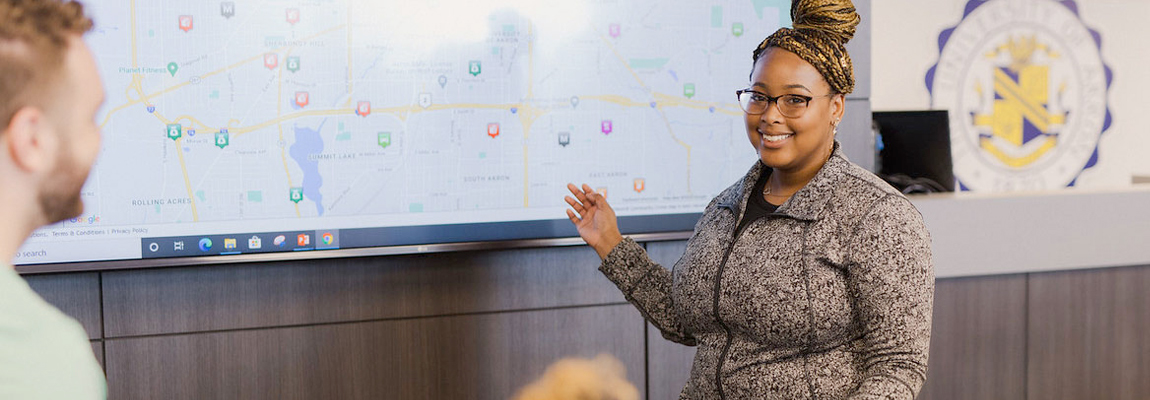 A woman standing in front of a projected screen giving a presentation to other people in the roon.